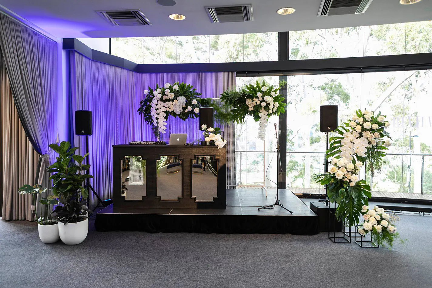 Wedding event setup with DJ booth, speakers on stands, microphone and floral arrangements around a small stage inside a bright venue
