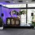Wedding event setup with DJ booth, speakers on stands, microphone and floral arrangements around a small stage inside a bright venue