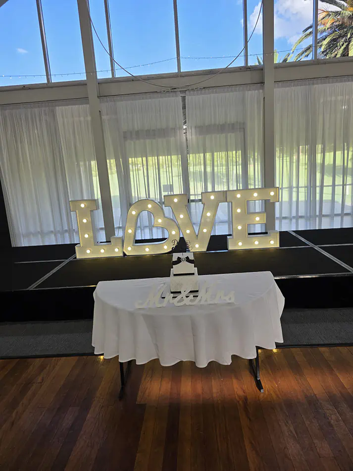 Wedding stage with white draping, illuminated LOVE letters and a cake table set in front on polished timber flooring