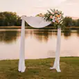 White wooden arbor styled with flowing white drapes and peach-toned roses, positioned by a lakeside at sunset for an elegant wedding ceremony backdrop.