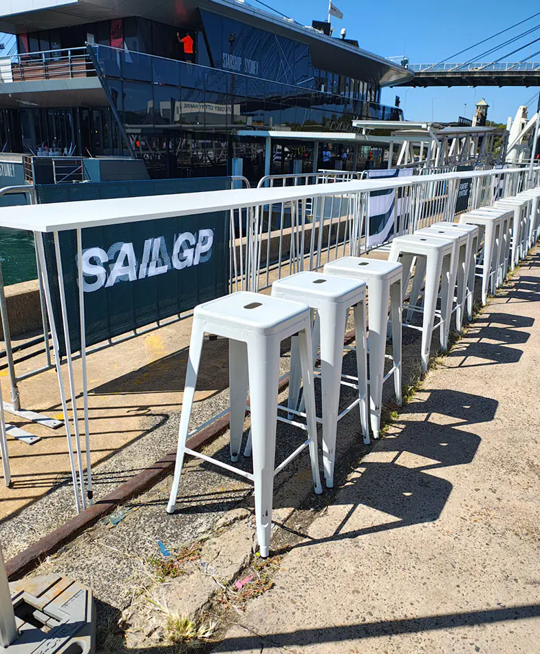 White bar stools lined along a high table overlooking a waterfront event area with building backdrop and outdoor seating arrangement