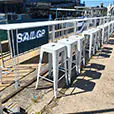 White bar stools lined along a high table overlooking a waterfront event area with building backdrop and outdoor seating arrangement