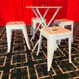 White metal bar table with four matching stools arranged on a geometric black carpet in front of a red curtain backdrop at an event venue.