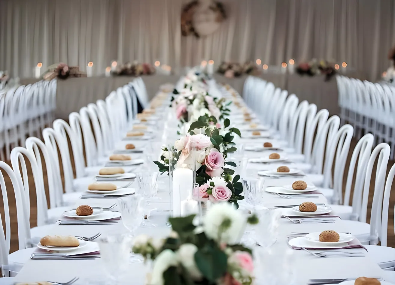 Rows of white bentwood chairs lining both sides of a long white-clothed reception table, decorated with a floral runner of pink and white roses, greenery, and white pillar candles inside a draped venue