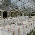 White bentwood chairs arranged around long rectangular tables dressed in white linen with blush napkins inside a clear-roof marquee, with globe pendant lights overhead and floral centrepieces in soft white and peach tones