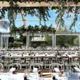 Rows of white bentwood chairs arranged along long rustic timber banquet tables inside a clear-roof marquee, set with wine glasses and white linen, with lush green and pink floral installations hanging overhead