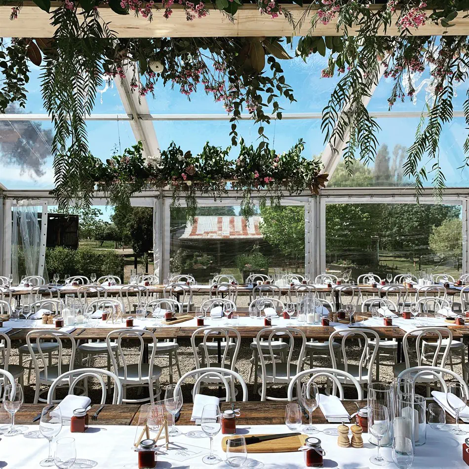 Rows of white bentwood chairs arranged along long rustic timber banquet tables inside a clear-roof marquee, set with wine glasses and white linen, with lush green and pink floral installations hanging overhead