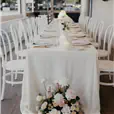 White bentwood chairs arranged along both sides of a long rectangular dining table dressed in a white linen tablecloth, with pillar candles, bud vases, and pink and white floral arrangements, set inside a bright semi-open venue