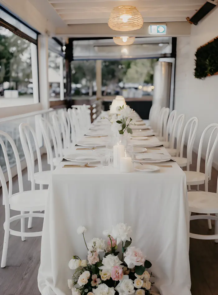 White bentwood chairs arranged along both sides of a long rectangular dining table dressed in a white linen tablecloth, with pillar candles, bud vases, and pink and white floral arrangements, set inside a bright semi-open venue