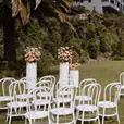 Rows of white bentwood chairs arranged on a manicured lawn at an outdoor wedding ceremony, with white floral pedestals marking the altar and a modern hillside building visible in the background