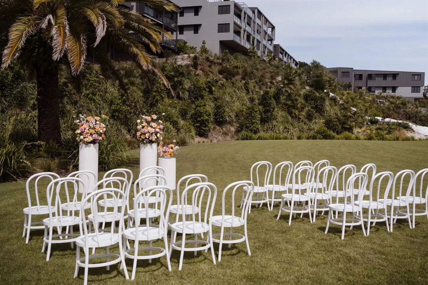 Rows of white bentwood chairs arranged on a manicured lawn at an outdoor wedding ceremony, with white floral pedestals marking the altar and a modern hillside building visible in the background