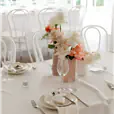 White bentwood chairs arranged around a round reception table set with white linen, wine glasses, and ceramic vases of white and peach flowers