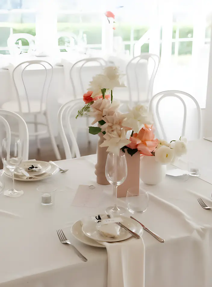 White bentwood chairs arranged around a round reception table set with white linen, wine glasses, and ceramic vases of white and peach flowers