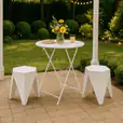 White bistro table styled outdoors with flowers and drinks, paired with white puzzle stools under a pergola with festoon lighting for a relaxed garden event setting.