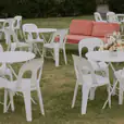 White bistro tables paired with matching white chairs arranged across a lawn for an outdoor event, with floral centrepieces and coral lounge seating in the background.