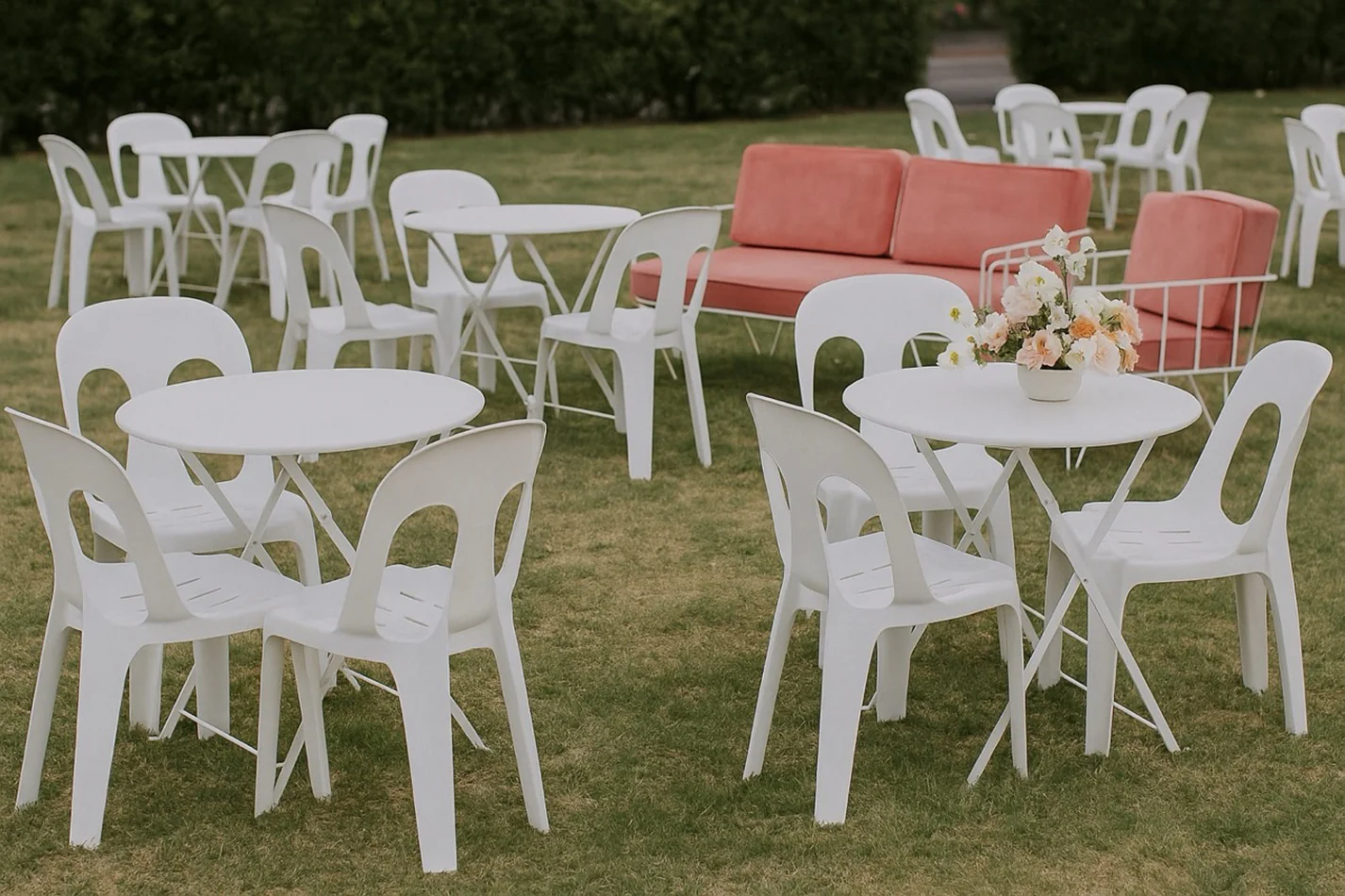 White bistro tables paired with matching white chairs arranged across a lawn for an outdoor event, with floral centrepieces and coral lounge seating in the background.