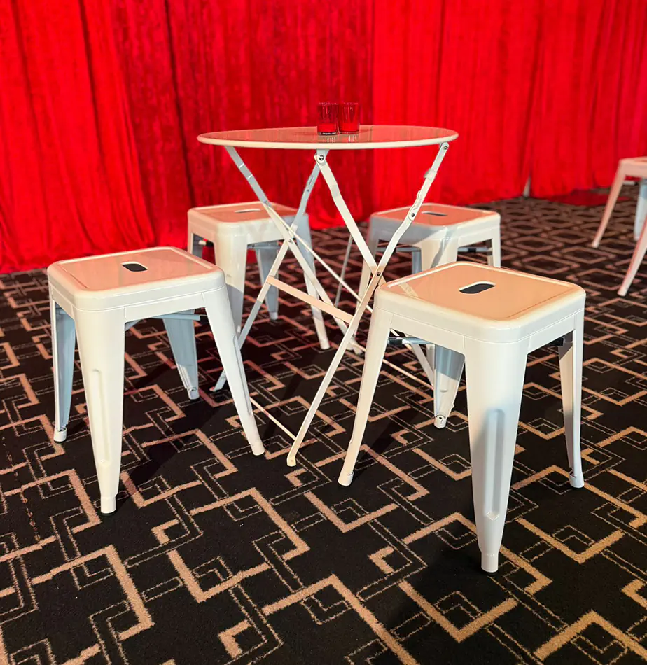 White cafe-style table with matching white metal stools positioned in front of red event draping on patterned carpet flooring