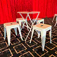White cafe-style table with matching white metal stools positioned in front of red event draping on patterned carpet flooring