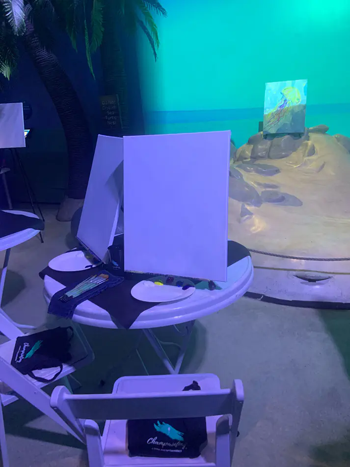 White café table set up for a paint and sip style activity with canvases, brushes, and art supplies in a themed indoor environment