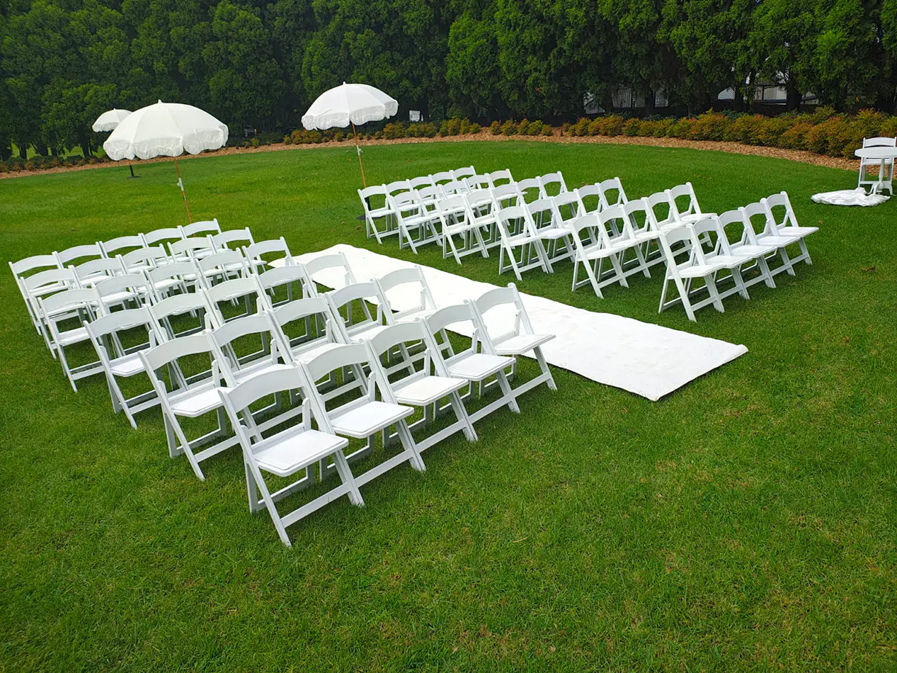 White folding ceremony chairs arranged on grass with a central aisle runner and umbrellas in an outdoor wedding-style setup