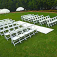 White folding ceremony chairs arranged on grass with a central aisle runner and umbrellas in an outdoor wedding-style setup
