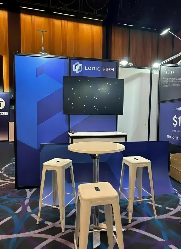 A white cocktail table paired with three white Tolix bar stools at a corporate exhibition booth, set up for a professional trade show or conference event.