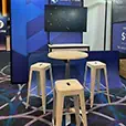 A white cocktail table paired with three white Tolix bar stools at a corporate exhibition booth, set up for a professional trade show or conference event.