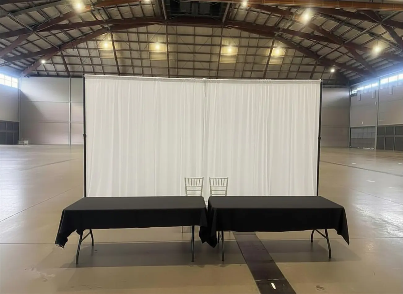 White draping behind two black-covered tables and chairs in a large exhibition or warehouse-style event space