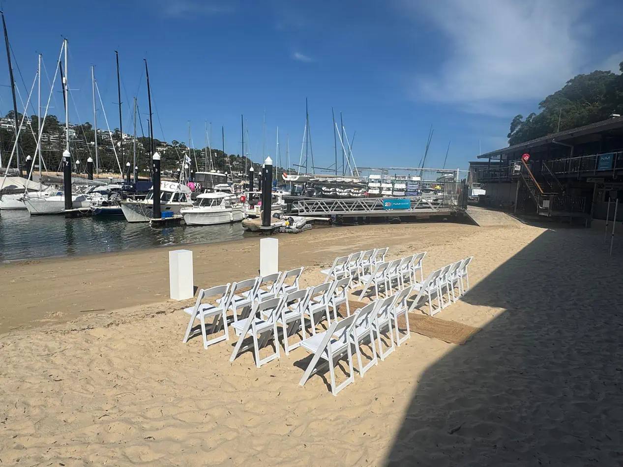 White folding chairs arranged on sand for a beachside ceremony near a marina with boats and waterfront backdrop