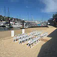 White folding chairs arranged on sand for a beachside ceremony near a marina with boats and waterfront backdrop