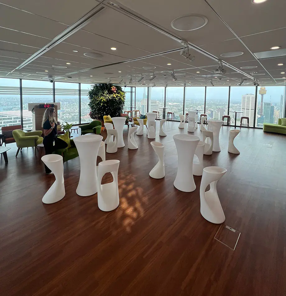 White illuminated stools and cocktail tables arranged in a modern indoor event space with timber flooring and city views through large windows