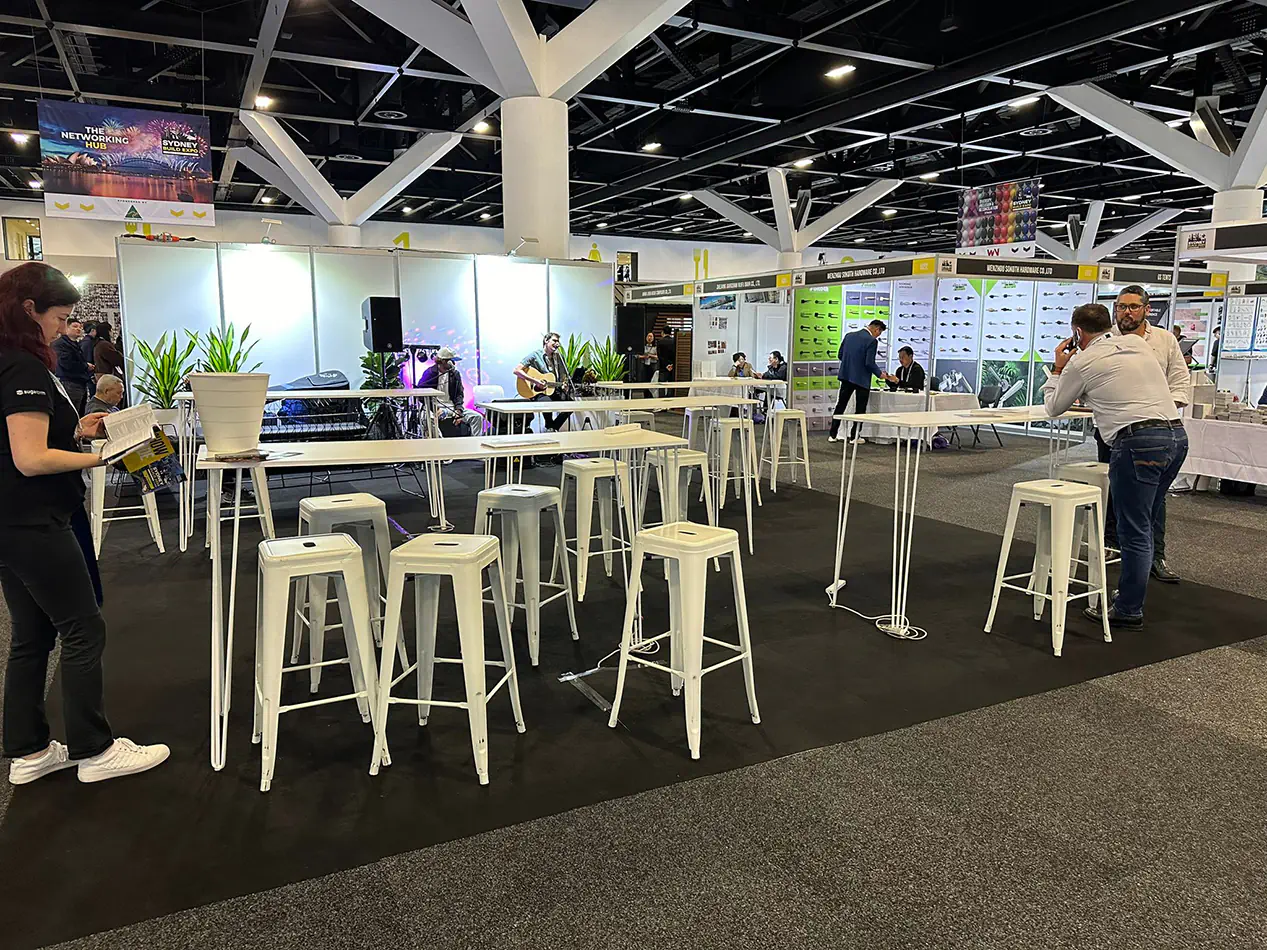 White hairpin bar tables paired with white Tolix stools in a large indoor expo event space with exhibition stands and overhead lighting