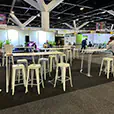 White hairpin bar tables paired with white Tolix stools in a large indoor expo event space with exhibition stands and overhead lighting