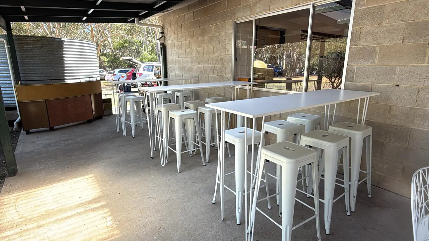 White hairpin bar tables with white Tolix stools arranged in a covered outdoor area beside a brick wall and glass doors