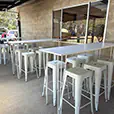 White hairpin bar tables with white Tolix stools arranged in a covered outdoor area beside a brick wall and glass doors