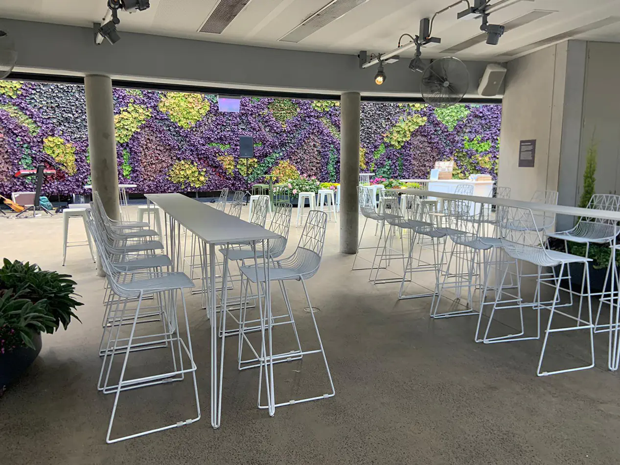 White hairpin bar tables with white wire chairs set in a modern indoor venue featuring a large green wall and open event layout