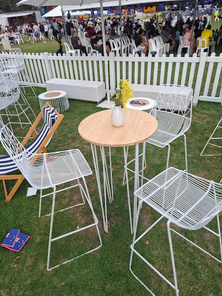 A white hairpin round cocktail table with a timber top styled with a small vase of yellow flowers, surrounded by white wire bar stools at an outdoor festival or lawn event.