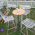 A white hairpin round cocktail table with a timber top styled with a small vase of yellow flowers, surrounded by white wire bar stools at an outdoor festival or lawn event.