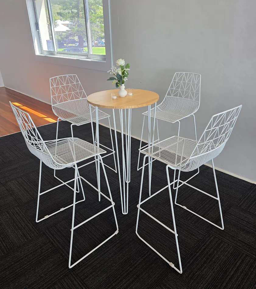 White round cocktail table with timber top and hairpin legs surrounded by white wire chairs in a clean indoor setting with natural light from large windows