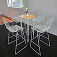 White round cocktail table with timber top and hairpin legs surrounded by white wire chairs in a clean indoor setting with natural light from large windows