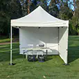 White pop up marquee with side walls set up on grass, with a serving table, chairs and weighted legs inside the sheltered space