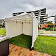 White pop up marquee with side walls set up on grass beside a paved courtyard and children's play area, with apartment buildings in the background