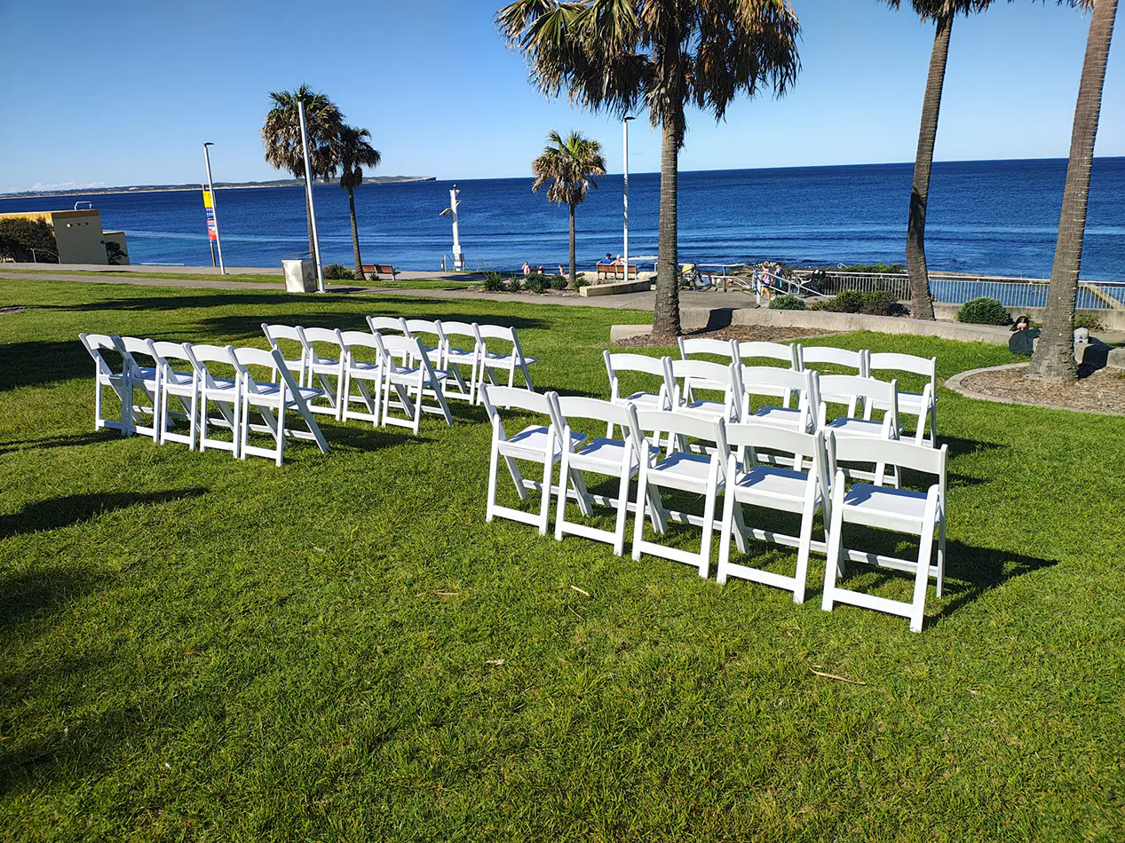 White padded folding chairs arranged on grass for a beachfront outdoor ceremony with ocean views and palm trees