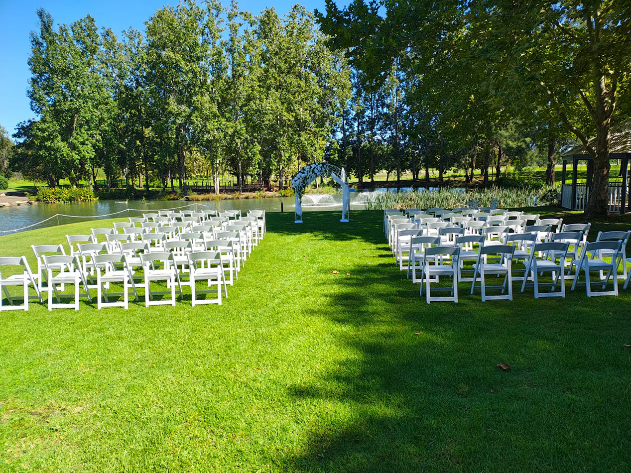 White padded folding chairs arranged in rows on grass for an outdoor wedding ceremony beside a lake with floral arbour