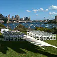 White padded folding Gladiator chairs arranged in rows for an outdoor wedding ceremony on a harbour lawn, with the Sydney Harbour Bridge and city skyline in the background.
