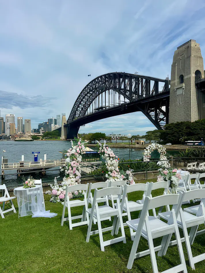 White padded folding Gladiator chairs set up for a waterfront wedding ceremony with a pink and white floral arch, overlooking Sydney Harbour Bridge.