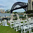 White padded folding Gladiator chairs set up for a waterfront wedding ceremony with a pink and white floral arch, overlooking Sydney Harbour Bridge.