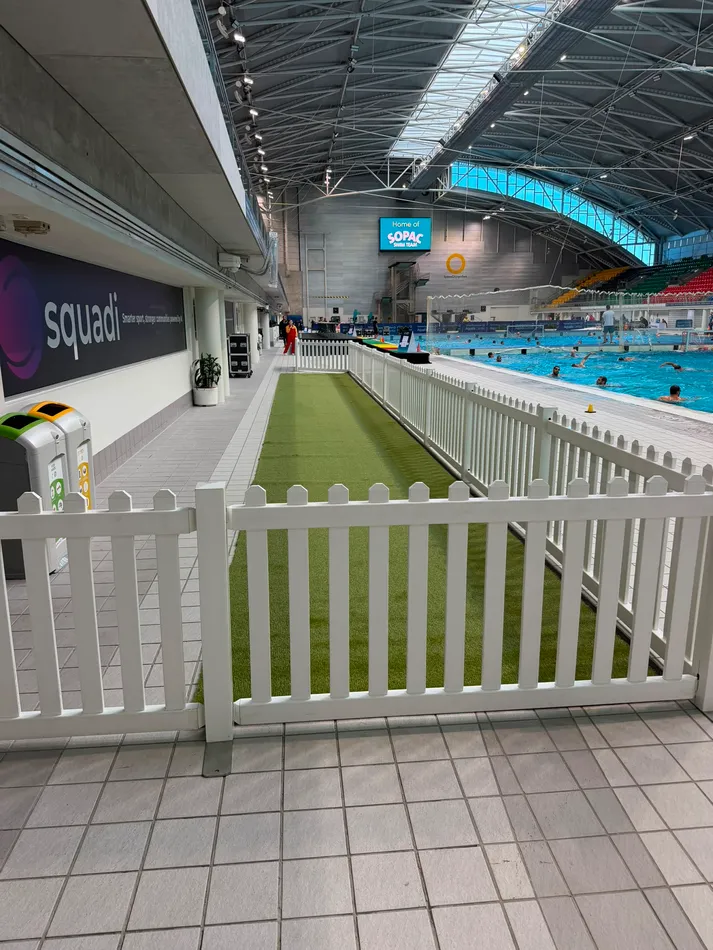 White picket fencing lining a green astro turf walkway beside the competition pool at an indoor aquatic centre