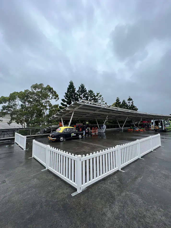 White picket fence panels forming a barrier around an outdoor covered area with vehicles and event setup on a wet surface