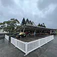 White picket fence panels forming a barrier around an outdoor covered area with vehicles and event setup on a wet surface